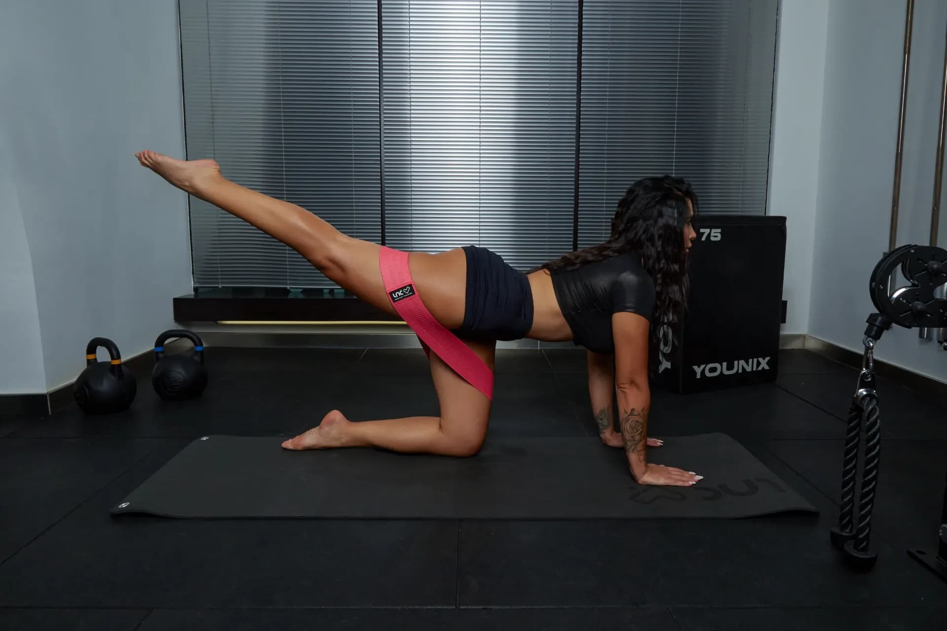 Un kickback a terra con elastico, eseguito su un tappetino accanto a due kettlebell e a un plyo box.